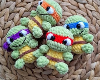 May include: Four handmade crochet turtle toys in green, yellow, and brown. Each turtle has a different colored headband: orange, blue, red, and purple. The turtles have black eyes and are arranged on a woven, light brown surface.