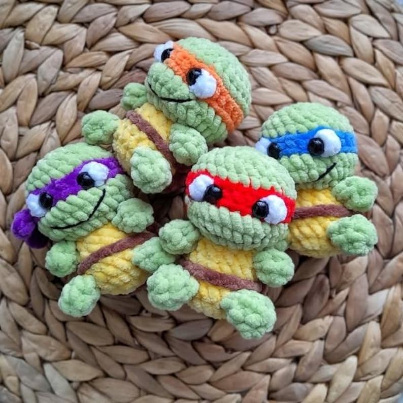 May include: Four handmade crochet turtle toys in green, yellow, and brown. Each turtle has a different colored headband: orange, blue, red, and purple. The turtles have black eyes and are arranged on a woven, light brown surface.