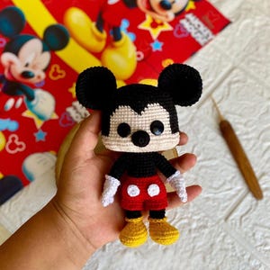 Pode incluir: Boneca Mickey Mouse de crochê feita à mão. A boneca tem orelhas pretas, corpo preto, calções vermelhos com botões brancos, sapatos amarelos e luvas brancas. O fundo é um tecido vermelho com ilustrações do Mickey Mouse.