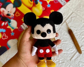 May include: A handmade crochet Mickey Mouse doll. The doll features black ears, a black body, red shorts with white buttons, yellow shoes, and white gloves. The background includes a red fabric with Mickey Mouse illustrations.