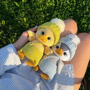 May include: Three crocheted chick toys, each wearing a hat with a white pom-pom and dungarees. One chick is yellow with a green hat, another is blue with a light blue hat, and the third is yellow with a light blue hat.