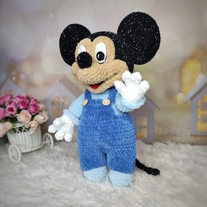 Puede incluir: Un peluche hecho a mano de un personaje de dibujos animados de rat&oacute;n. El rat&oacute;n tiene grandes orejas negras, una cara de color canela y lleva un peto azul con botones de madera. El juguete tiene guantes blancos y una cola negra. El fondo est&aacute; borroso con iluminaci&oacute;n suave.