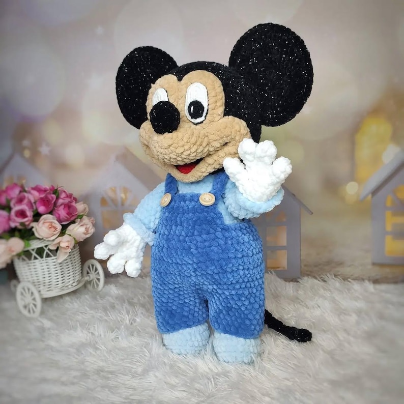 Puede incluir: Un peluche hecho a mano de un personaje de dibujos animados de rat&oacute;n. El rat&oacute;n tiene grandes orejas negras, una cara de color canela y lleva un peto azul con botones de madera. El juguete tiene guantes blancos y una cola negra. El fondo est&aacute; borroso con iluminaci&oacute;n suave.