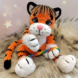 May include: A handmade orange and black crochet tiger plush toy with white paws and a pink nose. The tiger is holding a rainbow-coloured candy cane. The toy has black whiskers and is sitting on a furry surface.
