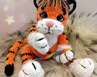May include: A handmade orange and black crochet tiger plush toy with white paws and a pink nose. The tiger is holding a rainbow-colored candy cane. The toy has black whiskers and is sitting on a furry surface.