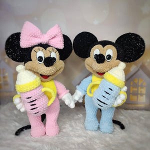 Może przedstawiać: Dwie pluszowe lalki Myszki Miki i Myszki Minnie, każda trzymająca butelkę dla dziecka. Myszka Minnie ma na sobie różowy strój i kokardę, a Myszka Miki jest w niebieskim. Oboje mają czarne uszy, opalone twarze i żółte akcenty.