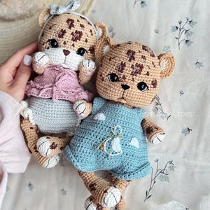 Op de afbeelding: Twee handgemaakte gehaakte luipaardpoppen. De ene pop draagt een roze jurk en een grijze strik, de andere een blauwe jurk. De poppen hebben bruine vlekken, bruine lichamen en witte accenten. De poppen liggen op een stof met bloemenpatroon.