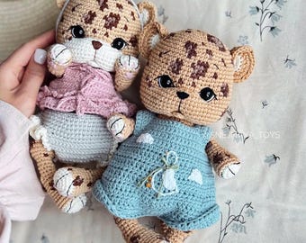 May include: Two handmade crochet leopard dolls. One doll wears a pink dress and a gray bow, the other wears a blue dress. The dolls have brown spots, tan bodies, and white accents. The dolls are lying on a floral patterned fabric.