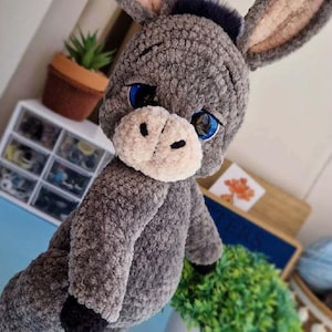 Op de afbeelding: Een handgemaakte grijze pluche ezel met grote blauwe ogen, een witte snuit en zwarte hoeven. De ezel heeft grote, slappe oren met roze binnenoren. Het pluche dier is een zacht, knuffelig speelgoed.
