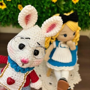 Puede incluir: Mu&ntilde;eco de conejo blanco tejido a ganchillo con gafas de montura met&aacute;lica, ropa roja y azul y un detalle en forma de coraz&oacute;n. Junto a &eacute;l, una mu&ntilde;eca de Alicia en el pa&iacute;s de las maravillas tejida a ganchillo con un vestido azul, un delantal blanco y un lazo negro.