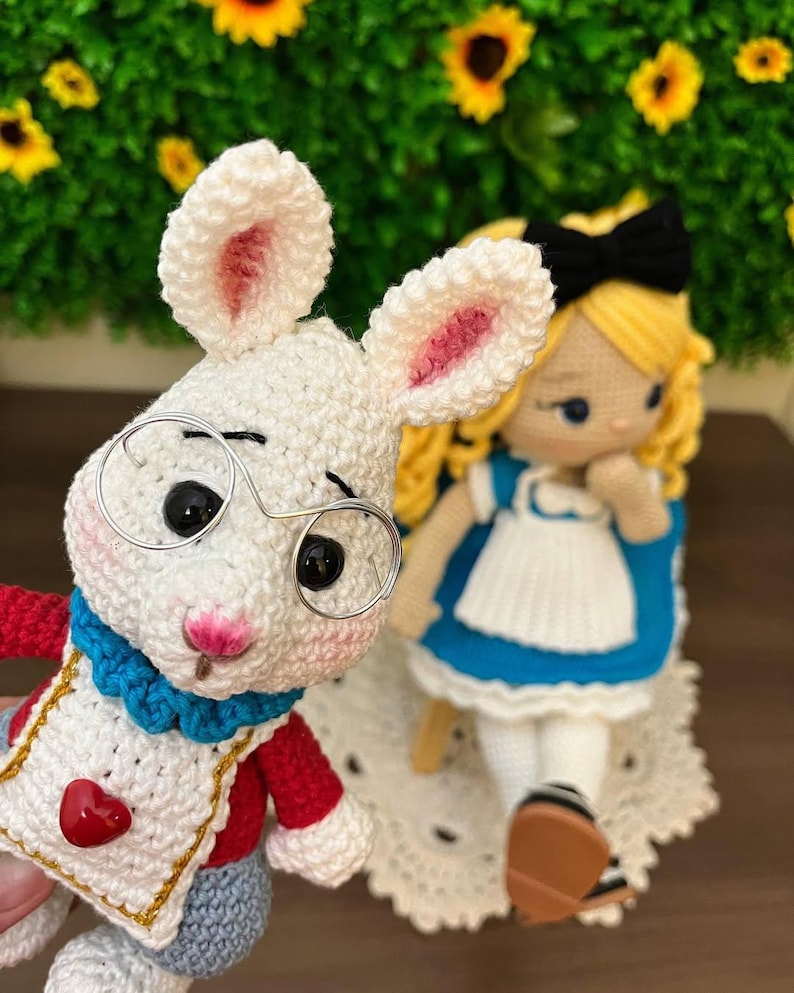 Puede incluir: Mu&ntilde;eco de conejo blanco tejido a ganchillo con gafas de montura met&aacute;lica, ropa roja y azul y un detalle en forma de coraz&oacute;n. Junto a &eacute;l, una mu&ntilde;eca de Alicia en el pa&iacute;s de las maravillas tejida a ganchillo con un vestido azul, un delantal blanco y un lazo negro.