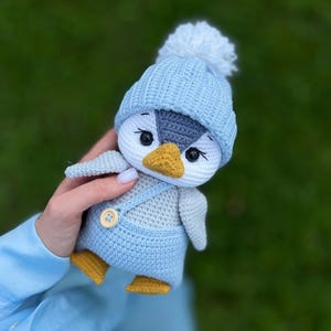 May include: A crocheted penguin toy, featuring a light blue hat with a white bobble, light blue dungarees, and yellow feet. The penguin has a white face, grey wings, and a yellow beak. The doll is held against a blurred green background.
