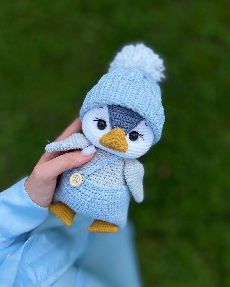 May include: A crocheted penguin toy, featuring a light blue hat with a white bobble, light blue dungarees, and yellow feet. The penguin has a white face, grey wings, and a yellow beak. The doll is held against a blurred green background.