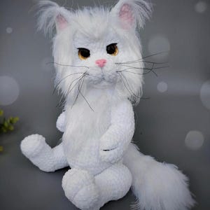 Może przedstawiać: Ręcznie robiona biała pluszowa zabawka w kształcie kota z puszystym ogonem, uszami i twarzą. Kot ma żółte oczy, różowy nos i długie czarne wąsy. Pluszak siedzi na szarej powierzchni.