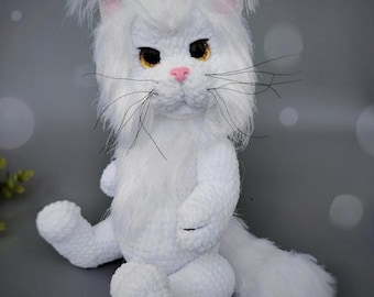 May include: A handmade white cat plush toy with a fluffy tail, ears, and face. The cat has yellow eyes, a pink nose, and long black whiskers. The plush is sitting on a gray surface.
