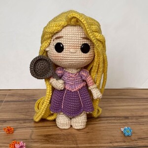 May include: A handmade crochet doll of a princess with long yellow hair, wearing a purple dress and holding a brown frying pan. The doll has large black eyes and is made with beige, pink, and purple yarn. Decorative crochet flowers are scattered around the doll.