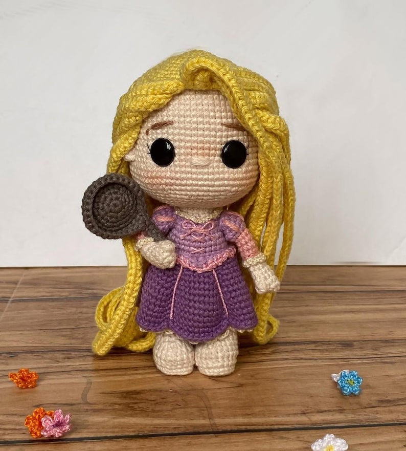 May include: A handmade crochet doll of a princess with long yellow hair, wearing a purple dress and holding a brown frying pan. The doll has large black eyes and is made with beige, pink, and purple yarn. Decorative crochet flowers are scattered around the doll.