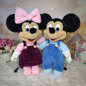 Puede incluir: Dos peluches de personajes de dibujos animados. Uno es un rat&oacute;n hembra con un lazo rosa, una blusa rosa y un vestido morado oscuro. El otro es un rat&oacute;n macho con un peto azul y una camisa azul claro. Ambos tienen orejas negras y caras color canela.