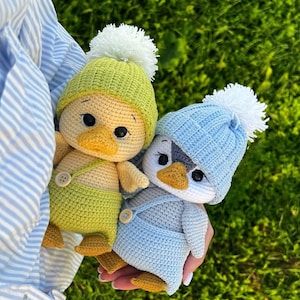 Puede incluir: Dos muñecos de ganchillo hechos a mano, uno amarillo y otro azul, se sostienen frente a un fondo verde. El muñeco amarillo lleva un gorro y un peto verdes, mientras que el muñeco azul lleva un gorro y un peto azules. Ambos muñecos tienen pompones blancos en sus gorros.