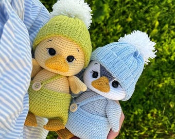 May include: Two handmade crochet dolls, one yellow and one blue, are held in front of a green background. The yellow doll wears a green hat and overalls, while the blue doll wears a blue hat and overalls. Both dolls have white pom-poms on their hats.