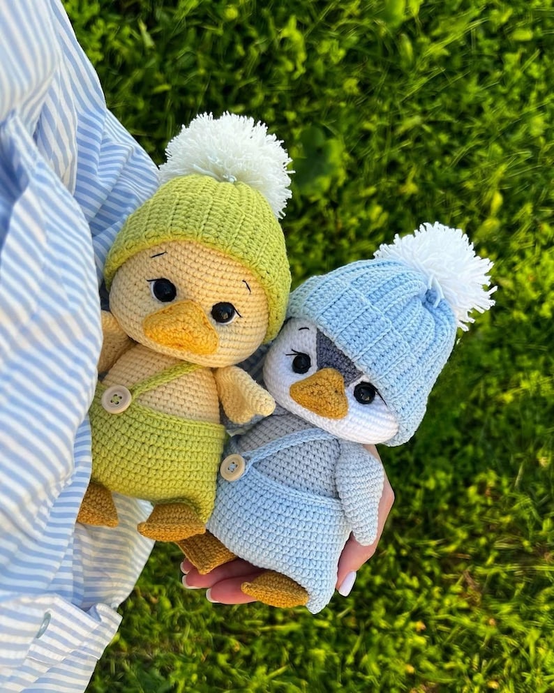 May include: Two handmade crochet dolls, one yellow and one blue, are held against a green background. The yellow doll wears a green hat and dungarees, while the blue doll wears a blue hat and dungarees. Both dolls have white pom-poms on their hats.