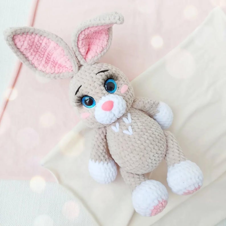 May include: A handmade plush bunny in soft beige yarn with pink accents. The bunny has large blue eyes with long eyelashes, a pink nose, and white paws. The ears are lined with pink. The bunny's chest features three small white hearts.