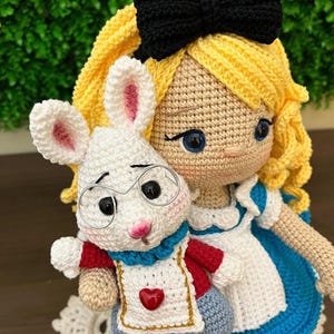 Puede incluir: Mu&ntilde;eca de ganchillo de una ni&ntilde;a con cabello rubio, un lazo negro y un vestido azul y blanco, sosteniendo un conejo blanco de ganchillo con gafas y un atuendo rojo y azul. El conejo tiene orejas internas rosas y un coraz&oacute;n rojo en su delantal.