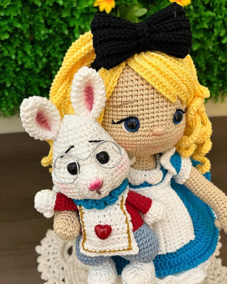 Puede incluir: Mu&ntilde;eca de ganchillo de una ni&ntilde;a con cabello rubio, un lazo negro y un vestido azul y blanco, sosteniendo un conejo blanco de ganchillo con gafas y un atuendo rojo y azul. El conejo tiene orejas internas rosas y un coraz&oacute;n rojo en su delantal.