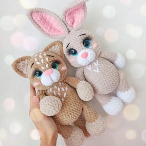 May include: Two handmade crochet stuffed animals: a tan bunny with pink inner ears and a tan cat with white spots. Both have blue eyes, pink noses, and white accents. The bunny has long ears, and the cat has small ears.