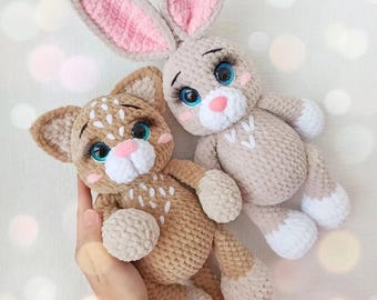 May include: Two handmade crochet stuffed animals: a tan bunny with pink inner ears and a tan cat with white spots. Both have blue eyes, pink noses, and white accents. The bunny has long ears, and the cat has small ears.