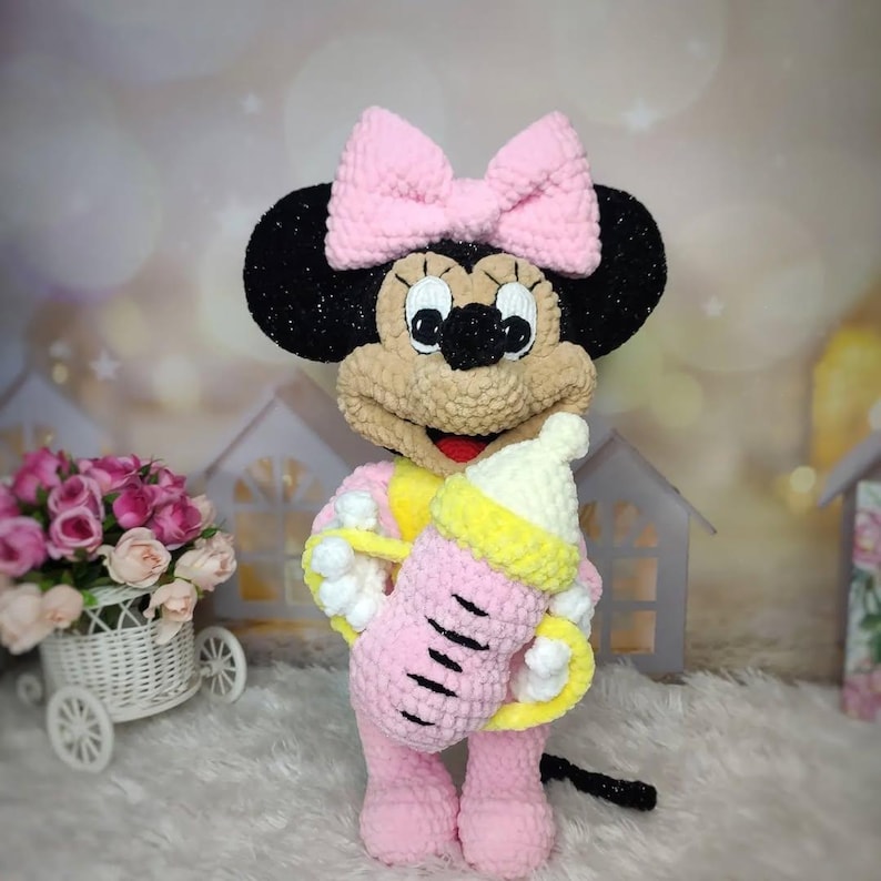 Puede incluir: Mu&ntilde;eca de Minnie Mouse de ganchillo hecha a mano en rosa, sosteniendo un biber&oacute;n rosa y amarillo. La mu&ntilde;eca tiene un gran lazo rosa, orejas negras y una cara bronceada. La mu&ntilde;eca lleva un atuendo rosa con detalles amarillos.