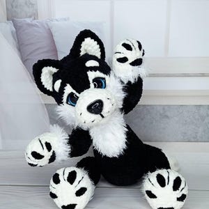 K&ouml;nnte beinhalten: Ein handgefertigtes H&auml;kel-Pl&uuml;schtier in Schwarz und Wei&szlig;, das einen Husky-Hund mit blauen Augen darstellt. Das Pl&uuml;schtier hat eine flauschige wei&szlig;e Brust und Pfoten und ist mit erhobenen Pfoten posiert. Das Spielzeug sitzt auf einer hellen Oberfl&auml;che.