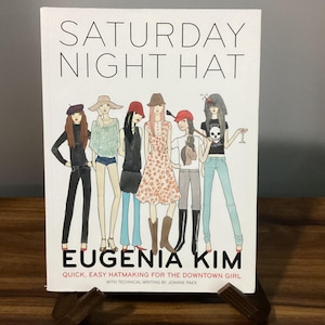 May include: A hardcover book titled SATURDAY NIGHT HAT by EUGENIA KIM. The white cover features a colorful illustration of six figures wearing various hats and outfits, with the subtitle QUICK, EASY HATMAKING FOR THE DOWNTOWN GIRL.
