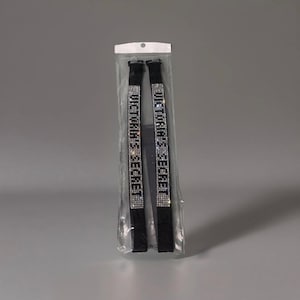 May include: Black elastic straps with the text "VICTORIA'S SECRET" spelled out in silver rhinestones. The straps are packaged in a clear plastic bag with a white header. The background is a solid gray.