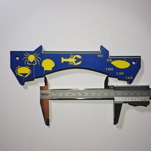 May include: A blue and yellow caliper tool with sea creature illustrations. The tool has a ruler with measurements in millimeters and a blue section with yellow crab, spider, shell, and lobster designs. Numbers are printed on the blue section.