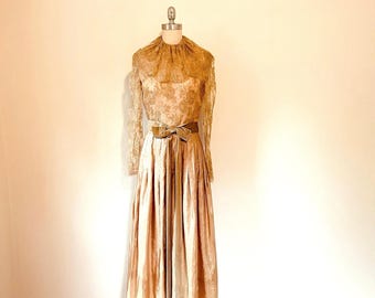 May include: A floor-length gold dress featuring a high neckline with a lace overlay, long lace sleeves, and a pleated skirt. A matching gold ribbon belt with a bow is tied at the waist, cinching the bodice above the flowing, full-length skirt.