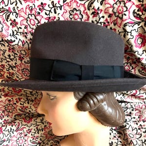Puede incluir: Un sombrero fedora de color marrón oscuro con una banda ancha de cinta de grosgrain negra y un pequeño lazo plano en el lateral. El sombrero tiene una copa estructurada con un pliegue central y un ala plana, sobre una cabeza de maniquí.
