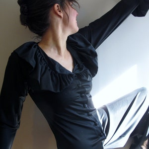 May include: A woman wearing a black long-sleeved blouse with a ruffled neckline and a silver skirt. The blouse has a button closure and the skirt is flowing.