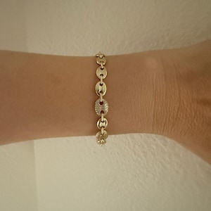 May include: A gold-colored bracelet with a chain-link design. The bracelet features oval links, some of which are adorned with small, sparkling stones. The bracelet is worn on a wrist, showcasing its design and style.