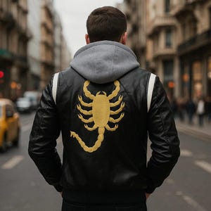 Könnte beinhalten: Schwarze Lederjacke mit einem goldenen Skorpion auf dem Rücken. Die Jacke hat eine graue Kapuze und weiße Streifen auf den Schultern. Die Person steht auf einer Stadtstraße.