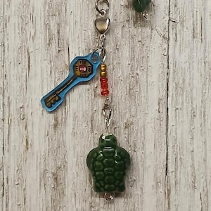 Puede incluir: Un llavero plateado con un dije de llave azul, cuentas rojas y doradas y un dije de tortuga verde. La llave tiene un diseño dorado. El llavero también tiene una colección de piedras de colores.