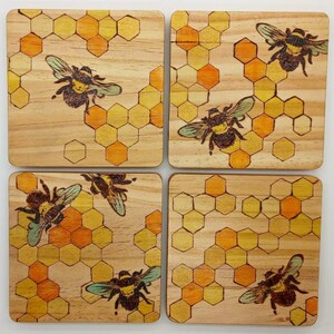 May include: Set of four square wooden coasters. Each coaster features a detailed illustration of a bee with green and brown wings, set against a honeycomb pattern in shades of yellow and orange. The coasters have a natural wood grain.