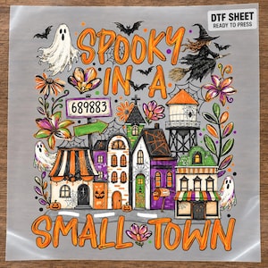 May include: A DTF sheet featuring a Halloween-themed graphic with the text SPOOKY IN A SMALL TOWN. The design includes a white ghost, a witch on a broomstick, bats, a water tower, colorful flowers, and a row of houses with the number 689883.