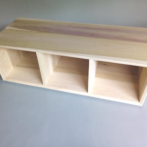 May include: A rectangular wooden storage unit featuring three open compartments. The top surface displays a natural wood grain with light tan and subtle purple-toned streaks, while the interior and sides show a lighter, pale beige wood finish.