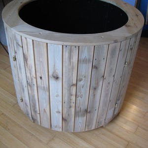 May include: A round wooden planter made from reclaimed wood planks. The planter has a black liner inside and is sitting on a wooden floor.