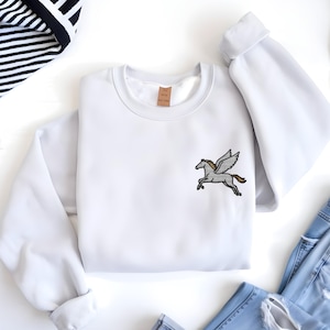 May include: A white crewneck sweatshirt featuring an embroidered Pegasus design on the front. The Pegasus is depicted in gray with tan and black accents. The sweatshirt is made of a soft material and is suitable for casual wear.