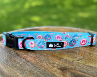 May include: A light blue dog collar featuring a repeating pattern of donuts in pink, blue, and brown. The collar has a black plastic buckle and a black metal D-ring. The brand name "Lulu Grazer" is displayed on a black label.