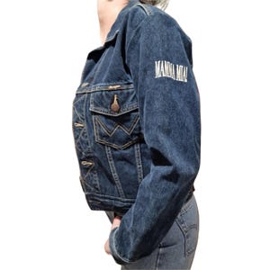 Puede incluir: Chaqueta vaquera azul oscuro con el texto "MAMMA MIA!" bordado en la manga. La chaqueta tiene cierre de botones, dos bolsillos en el pecho y un logotipo clásico de Wrangler en el bolsillo.
