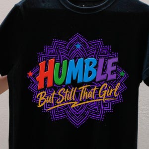 May include: A black short-sleeve t-shirt featuring a colorful graphic design with the text "HUMBLE But Still That Girl" in a bold, multi-colored font. The text is centered over a purple, intricate, mandala-style pattern with small stars.