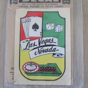 May include: A vintage decal featuring a Las Vegas theme. The design includes playing cards, dice, and a roulette wheel, with the words "Las Vegas Nevada" prominently displayed. The decal is in a clear plastic sleeve.